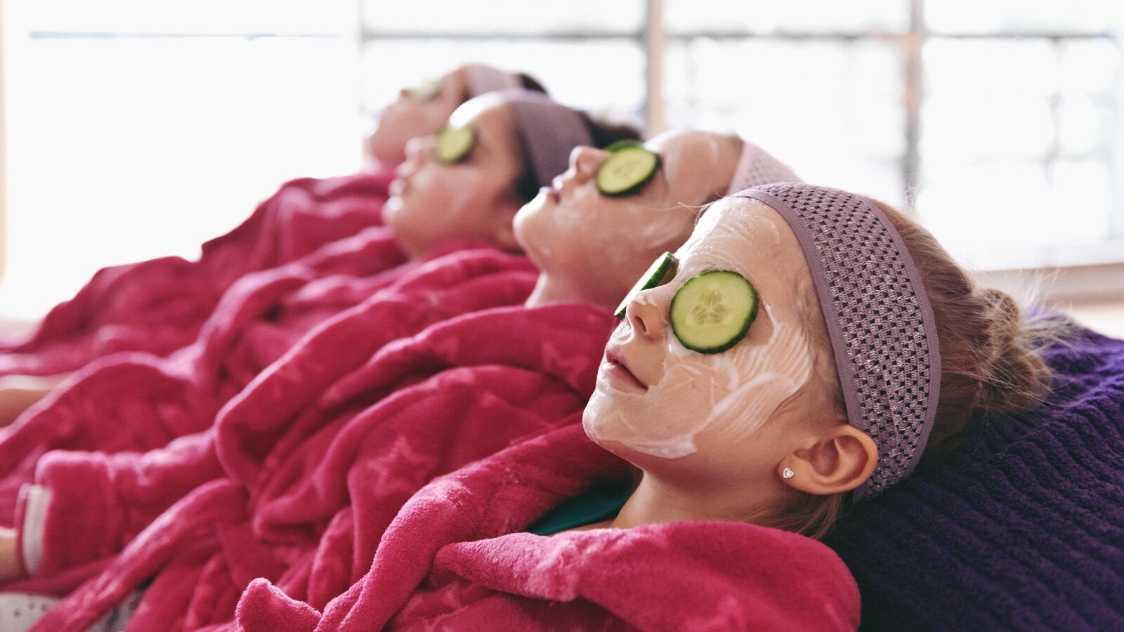 Des petites filles en peignoir ont un masque sur le visage ainsi que des concombres sur les yeux.