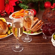 Des plats tels qu'une dinde, des carottes, des choux de Bruxelles, un jambon et une sauce aux canneberges ainsi que des verres de mousseux sont disposés sur une table pour Noël. 