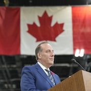 Un homme en complet est debout devant un drapeau canadien.