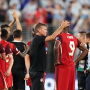 Dans un stade, un entraîneur console un joueur, devant d'autres joueurs qui applaudissent.
