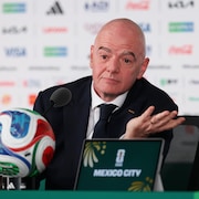 Un homme chauve lors d'une conf&eacute;rence de presse. Il est assis &agrave; une table sur laquelle est pos&eacute; un ballon de soccer.