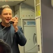 Debout dans un avion, Pierre Poilievre s'adresse aux passagers avec le système de communication de l'appareil.