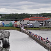 Des gens habillés aux couleurs de l’Acadie marchent sur une passerelle en direction d'une petite île.