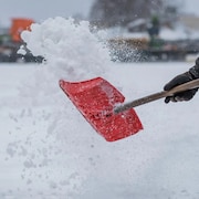 Une personne soulève une bonne quantité de neige avec une pelle, sur un grand terrain, à l'extérieur.