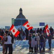 Des manifestants devant l'Assemblée législative, en hiver. Plusieurs portent des drapeaux du Canada.