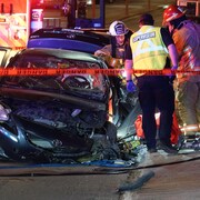 Des premiers répondants se tiennent près d'une voiture lourdement endommagée.
