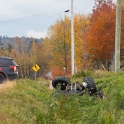 Un VTT renversé dans un fossé sur le bord d'une route avec un véhicule de police à l'arrière-plan.