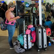 Dans un aéroport, une femme debout à côté de plusieurs valises à roulettes regarde son téléphone cellulaire, et trois enfants sont assis par terre.