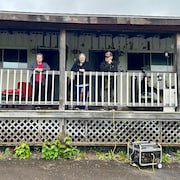 Trois personnes accotées à la balustrade et une personne assise sur la véranda d'un bâtiment en décrépitude. Une rallonge électrique raccorde la véranda à une génératrice posée devant, près d'un barbecue et d'une bonbonne de propane.
