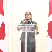 Amira Elghawaby entourée de deux drapeaux canadiens parle au micro en conférence de presse.