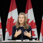 Chrystia Freeland, assise, s'exprime devant quatre drapeaux canadiens.