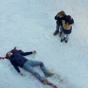 Dans un décor enneigé, deux personnes se tiennent debout devant un homme mort sur le sol.
