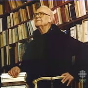 Le père Anselme Chiasson appuyé sur une pile de livres dans une bibliothèque. 
