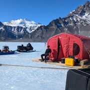 Un homme prend un bain de soleil près d'une tente rouge installée sur un lac glacé.