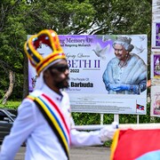 Un militaire caribéen en uniforme de cérémonie marche devant une affiche pour la mémoire d'Élisabeth II.