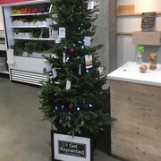 Un grand sapin décoré avec des lumières de Noël et des cartes de dons est installé dans un magasin. En arrière-plan, il y a un étalage avec des légumes frais.