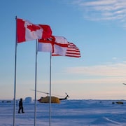 D'immenses drapeaux du Canada et des États-Unis, de même que le pavillon britannique White Enseign flottent au vent près de deux hélicoptères au sol dans l'Arctique.