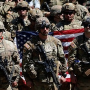 Des soldats américains armés devant un drapeau des États-Unis tenu par d'autres soldats.