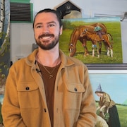 Josh Harmack sourit devant un mur où figurent plusieurs de ses toiles. 