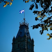 Plan rapproché et en contre-plongée de la tour de l’hôtel du parlement, au sommet duquel flotte un drapeau du Québec. À l’avant-plan, les branches d’un arbre feuillu.