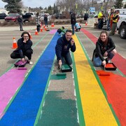Trois personnes accroupies peignent les couleurs de la fierté sur une chaussée.