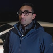 Un homme portant des lunettes devant sa voiture.