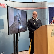 Le surintendant Glenn Sells, en charge de l'équipe intégrée de la sécurité nationale de la GRC, est au podium avec derrière lui un écran montrant la photo d'un homme tenant une arme.