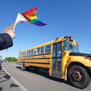 Une main brandit un drapeau LGBTQ+ le long d’une route où un autobus scolaire se déplace.