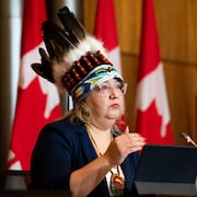 Une femme porte une coiffe traditionnelle autochtone et parle devant un micro.