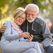 Assis sur un banc de parc, un couple de retraités s'amuse en consultant ensemble un même téléphone cellulaire.