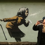 Une femme se prend en photo devant une murale de l'artiste de rue britannique Banksy illustrant une personne âgée qui éternue.