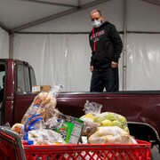 Des employés d'une banque alimentaire préparent une distribution. Une femme pousse un panier d'épicerie remplie de nourriture vers une camionnette. Un homme est debout dans la boîte du pickup.