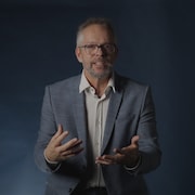 Un homme est assis sur un tabouret devant un mur bleu dans un studio.