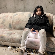Une personne en manteau d'hiver est assise sur un vieux sofa décrépit, entouré de déchets, dans un bâtiment abandonné. 