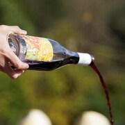 Du vin rouge coule d'une bouteille de vin à l'horizontale tenue par la main d'un homme qui porte une chemise blanche et un veston noire.