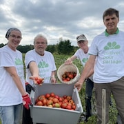 Quatre personnes avec un chandail blanc avec l'inscription Cultiver pour partager se tiennent debout dans un champ avec dans leurs mains des tomates.