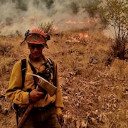 Benson Munch debout dans un champ. Derrière lui, on voit des flammes et de la fumée. Il porte un habit de pompier avec une hache à la main (été 2023).