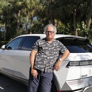 Bernard Pélissier devant sa voiture électrique stationnée près de palmiers.