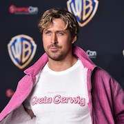 Ryan Gosling pose sur le tapis rouge, avec un manteau rose qu'il tient ouvert. Sur son t-shirt, on peut lire en anglais «De la réalisatrice Greta Gerwig». 