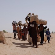 Des gens marchent dans le désert en portant des sacs en jute sur la tête.