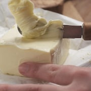 Gros plan sur des mains qui tiennent une brique de beurre et tartinent à l'aide d'un couteau à beurre. 
