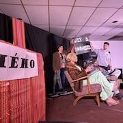 Sur une scène, deux comédiens sont assis sur des chaises, tandis que deux autres tiennent une pancarte derrière eux. 