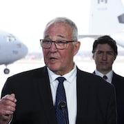 Bill Blair parle devant un micro avec Justin Trudeau en arrière-plan, le 8 avril 2024, en Ontario.