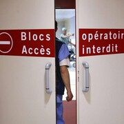 Deux portes blanches sont entrouvertes, permettant d'apercevoir deux employés de l'hôpital. Sur les portes, on peut lire : Blocs opératoires, accès interdit.