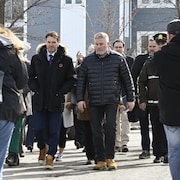 Deux hommes marchent côte à côte dans une rue ensoleillée, entourés de passants et de journalistes.