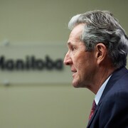 Brian Pallister est vu de profil, avec en arrière-plan une affiche montrant le logo du Manitoba. 