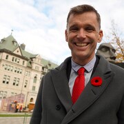 Bruno Marchand sourie pour la caméra, devant l'hôtel de Ville de Québec. Il porte un manteau gris avec une cravate rouge.