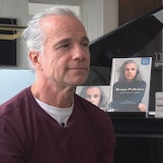Bruno Pelletier est assis devant un piano. Son album et son récit biographiques sont déposés sur le piano. L'artiste regarde la journaliste. 