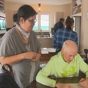 Une personne assis à une table est en train d'écrire sur une feuille alors qu'une autre personne debout est tournée vers elle. 