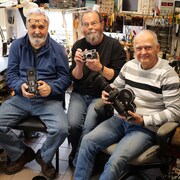 Trois hommes souriants aux cheveux gris tiennent des appareils-photos d'autrefois devant un établi couvert de fils et d'outils. 